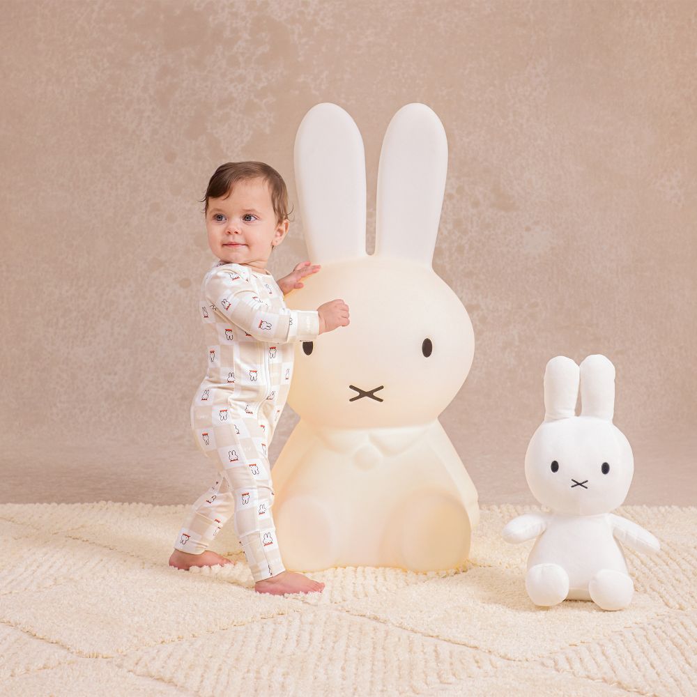 A baby in a Miffy print babygro by Love to Dream stood beside a large Miffy rabbit