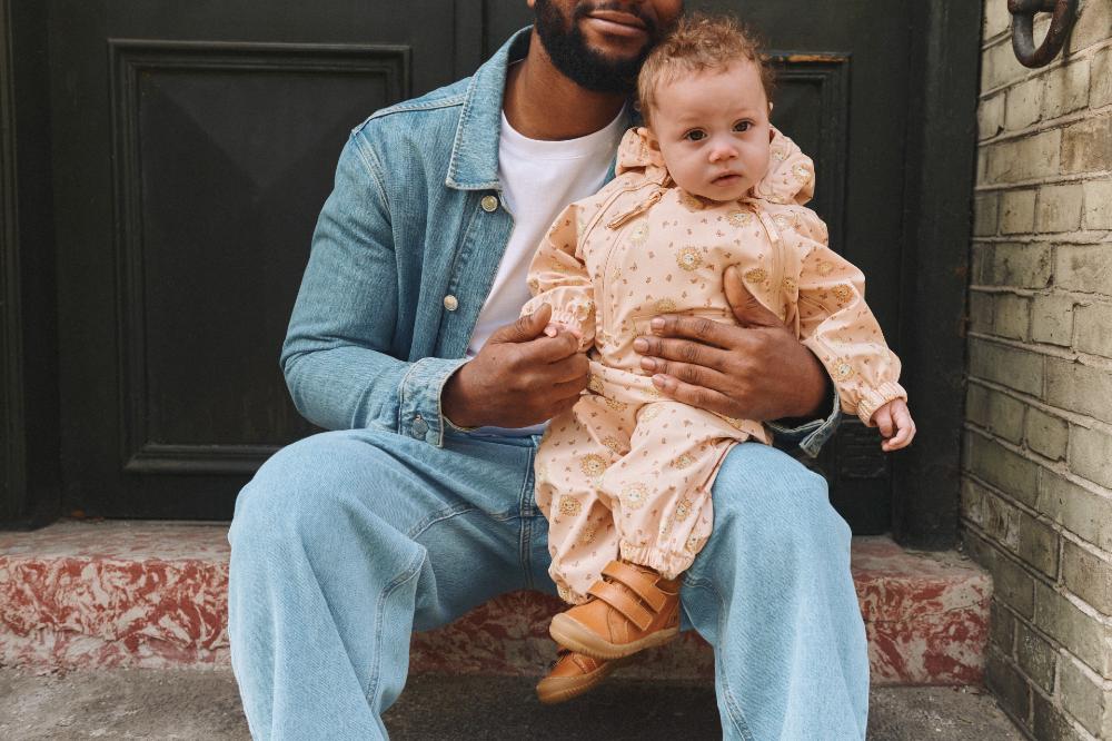 A man sat outside holding a baby wearing an all in one suit by Wheat