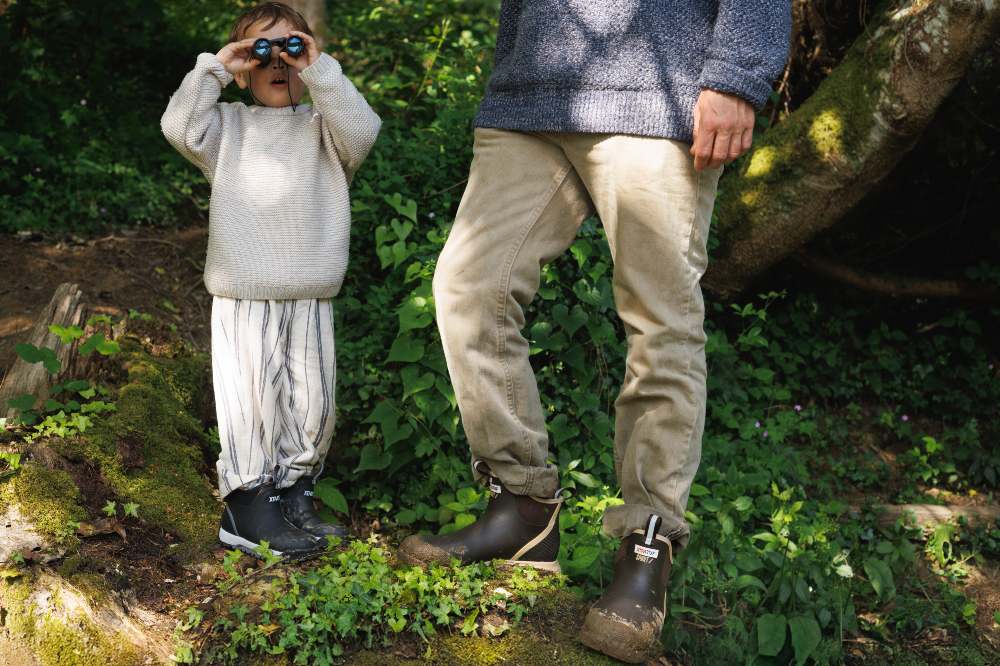 A child in a wood looking through binoculars stood beside an adult and both wearing XTRATUF waterproof boots