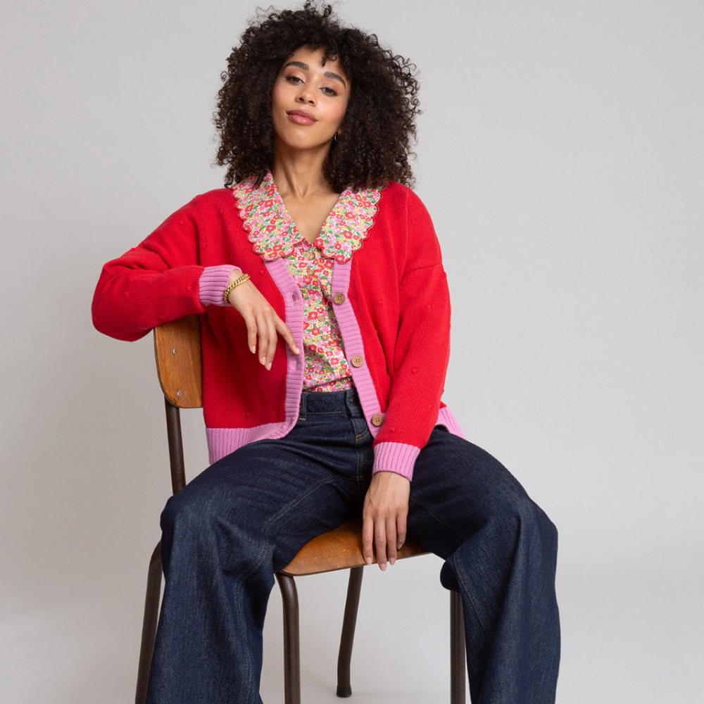 A woman sat in a chair wearing a frill collar blouse and red and pink cardigan from Kite Clothing's matching family Christmas collection