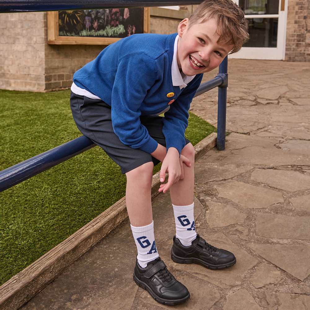 A school boy sat on a railing outside a school wearing a blue sweatshirt, grey shorts and black school shoes by Start-Rite 