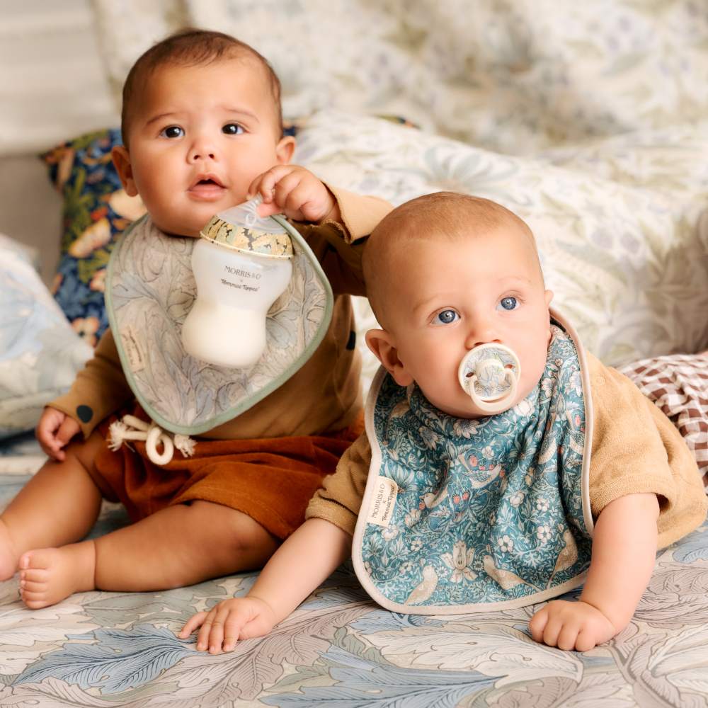 Two babies sat together on a bed wearing Tommee Tippee bibs