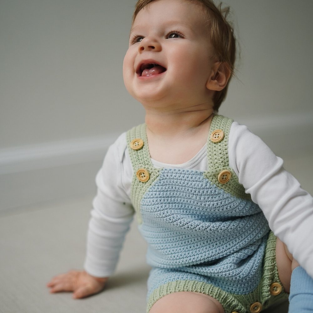 A smiling baby sat on the floor wearing a white top and a pale blue knitted playsuit