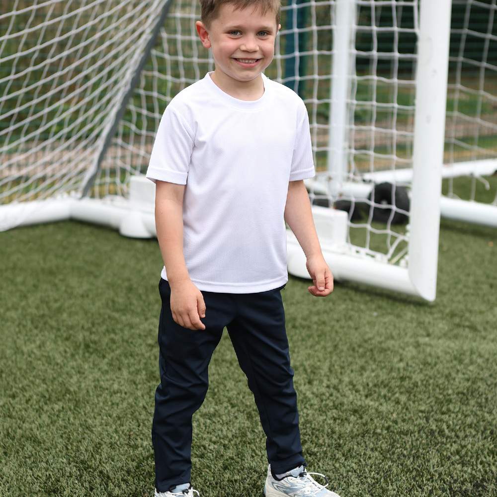 A young child in black training pants and a white T-shirt stood outside in front of a goal net 