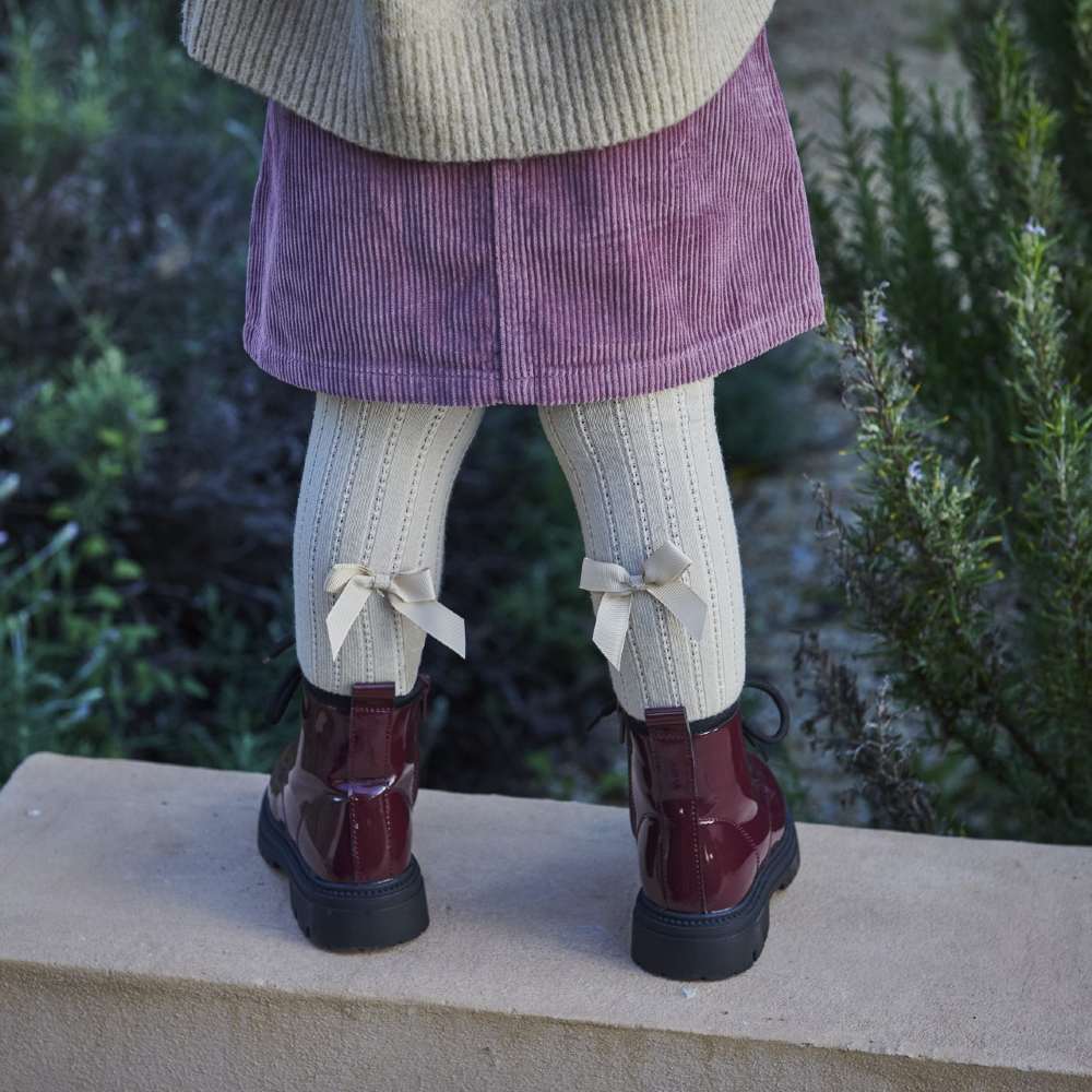 The back of a child shown stood outside on a wall wearing a purple skirt, knee socks with bows on the back and a boots