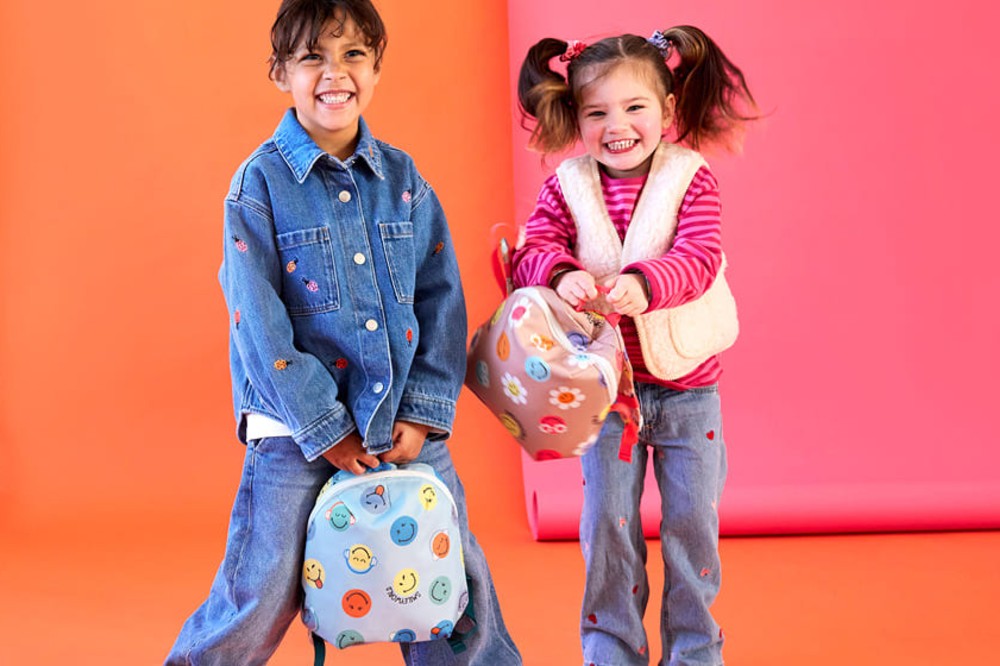 Two children stood in front of a pink and orange background holding rucksacks from the LÄSSIG x SmileyWorld® collection