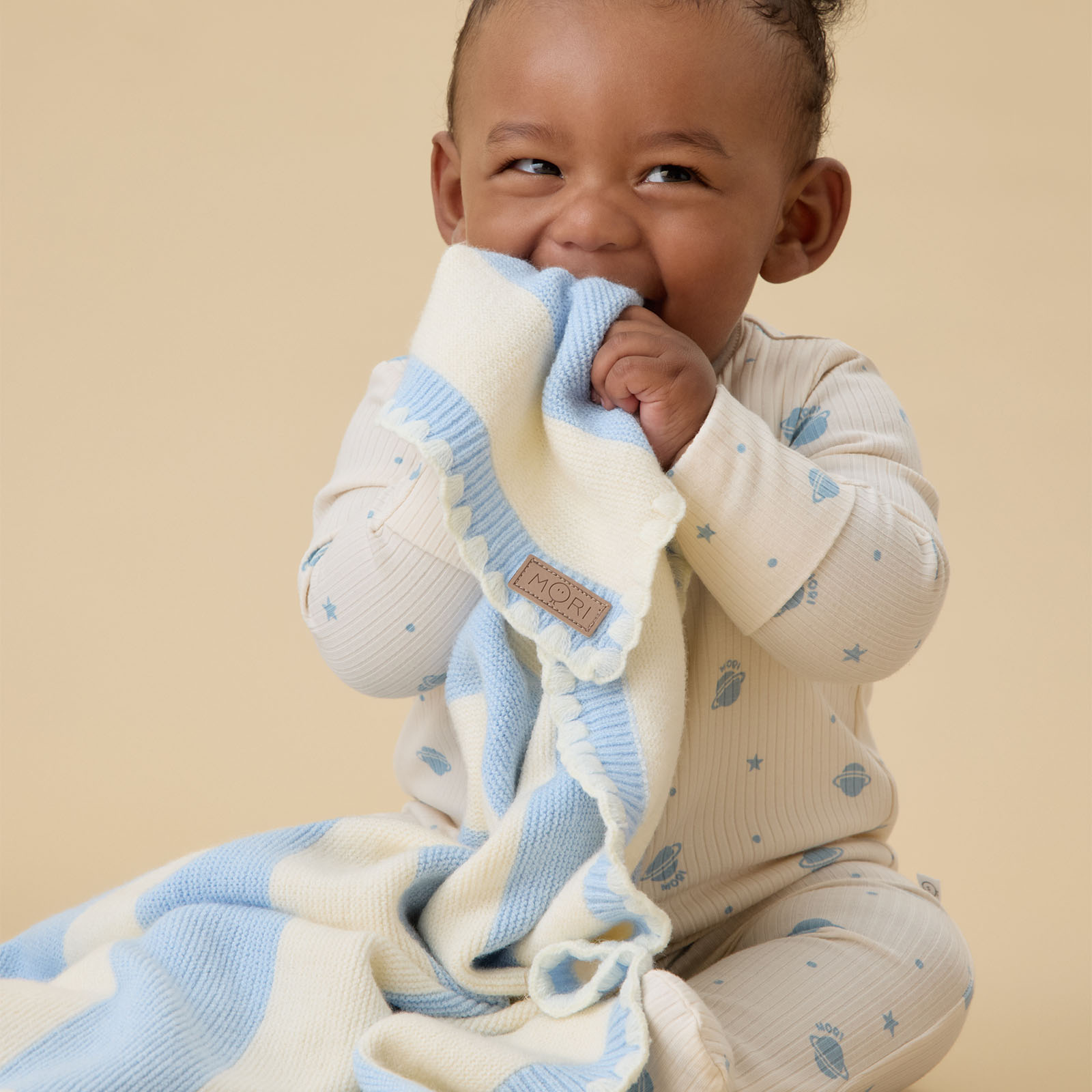 A baby sat on the floor holding a blue and white MORI baby blanket and wearing a matching babygro 