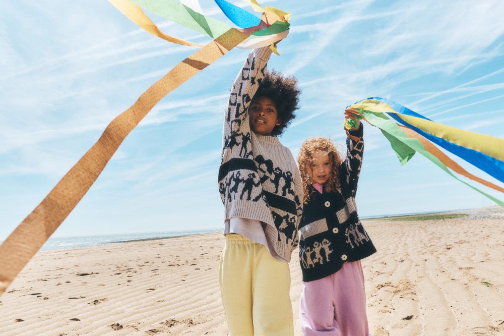 Two children on a beach holding ribbons and wearing knitwear by Talou
